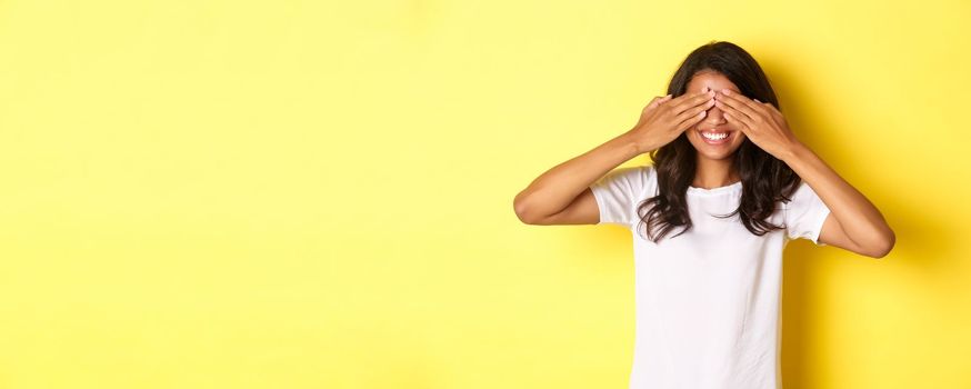 Image of excited african-american girl waiting for surprise, smiling and covering eyes with hands, standing over yellow background.