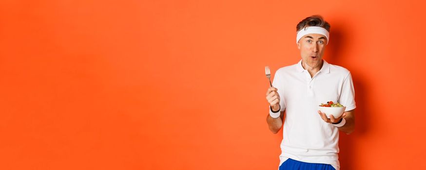 Concept of sport, fitness and lifestyle. Portrait of joyful middle-aged guy in workout uniform, holding fork and salad, eating healthy, standing over orange background.