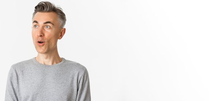 Close-up of handsome, middle-aged guy with gray hair, open mouth and looking amazed at upper left corner, standing against white background.