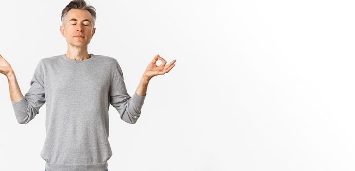 Portrait of calm and relaxed middle-aged man, breathing air freely, meditating with eyes closed and hands spread sideways, practice yoga over white background.