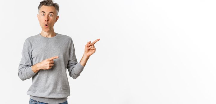 Amazed handsome guy in grey sweater, pointing fingers right and looking curious, askign question about product, standing over white background.