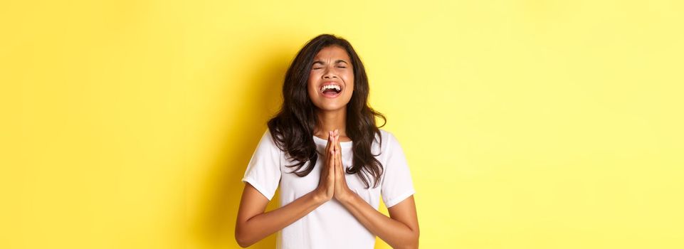 Portrait of young african-american girl pleading, begging for help with hands pressed together on chest, standing in white t-shirt over yellow background.