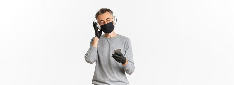 Concept of covid-19, social distancing and lifestyle. Image of middle-aged man in medical mask, gloves and glasses, listening music in headphones and holding smartphone.