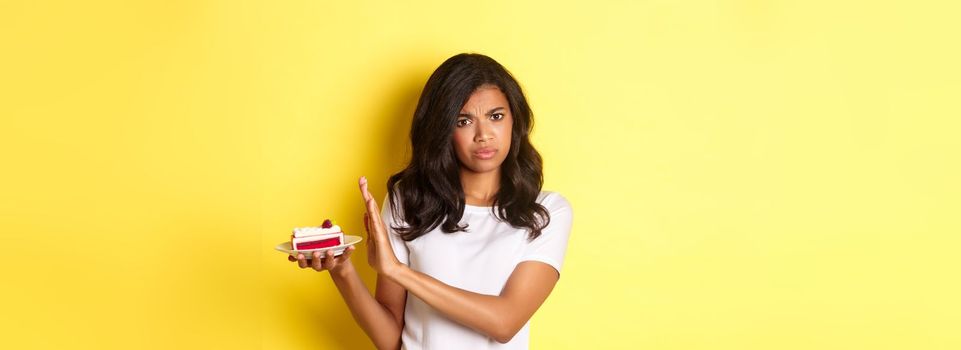 Image of displeased african-american girl decline to eat a cake, standing over yellow background.