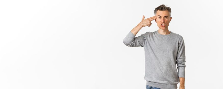 Image of annoyed and fed up middle-aged man showing finger gun sign near head, shooting himself with pissed-off face, standing tired over white background.