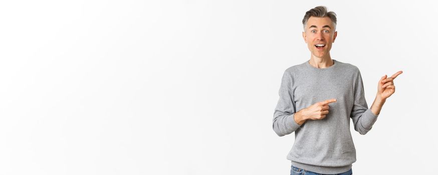 Image of surprised handsome middle-aged man, smiling and pointing fingers right at logo or promo banner, standing amazed over white background.