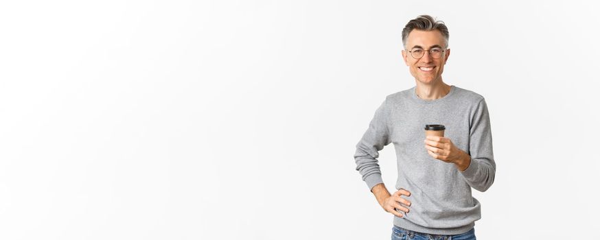 Image of confident and handsome middle aged man, wearing glasses and grey sweater, smiling happy and drinking coffee from takeaway cup, standing satisfied over white background.