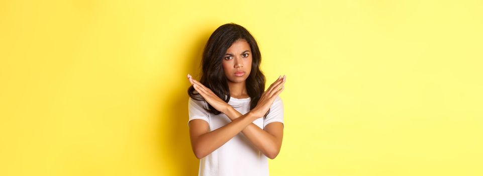 Image of disappointed and angry african-american woman, telling no, showing stop sign and frowning, prohibit and disagree with you, standing over yellow background.