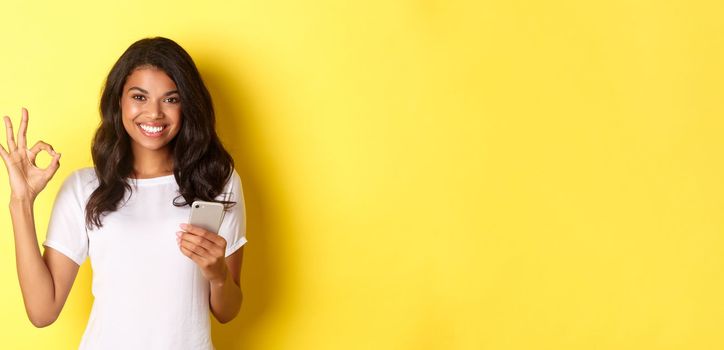 Image of good-looking african-american woman, showing okay sign with confidence, holding smartphone, standing over yellow background.