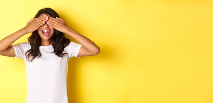 Image of excited african-american girl waiting for surprise, smiling and covering eyes with hands, standing over yellow background.