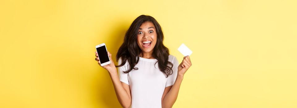 Image of excited african-american female model, showing smartphone screen and credit card, standing over yellow background.