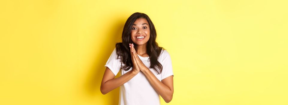 Image of happy and pleased african-american girl, clap hands and looking excited, thanking for something, standing over yellow background.