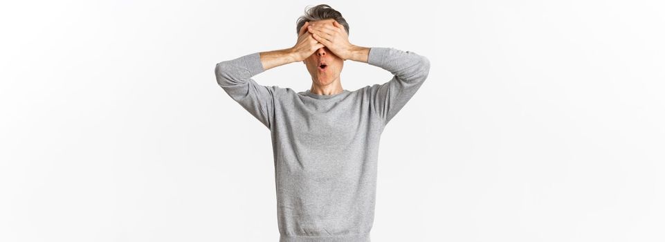 Image of excited middle-aged guy in grey sweater, cover eyes with hands and looking amazed while waiting for surprise, playing hide-n-seek, standing over white background.