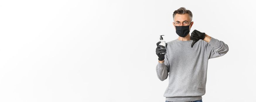 Concept of coronavirus, lifestyle and quarantine. Portrait of middle-aged man in medical mask and gloves do not recommend antiseptic, showing hand sanitizer and thumbs-down, white background.