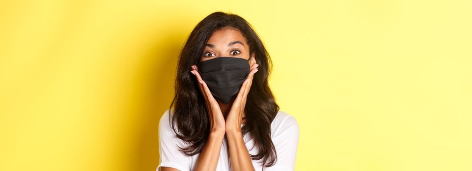 Concept of coronavirus, pandemic and lifestyle. Close-up of amazed african-american girl in black face mask, looking wondered at something awesome, yellow background.