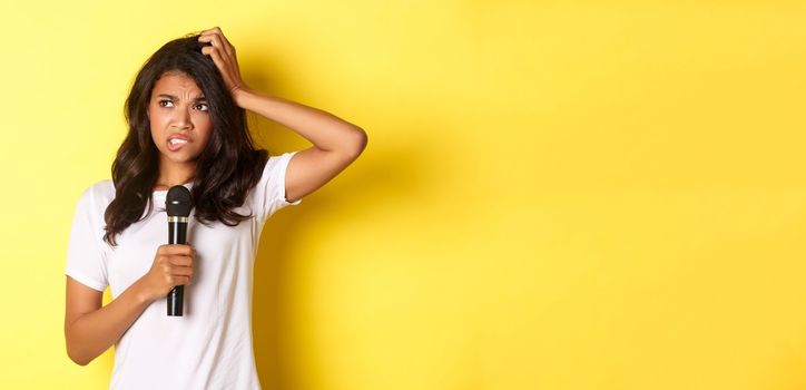 Image of indecisive african-american girl, holding microphone and looking hesitant, standing over yellow background.