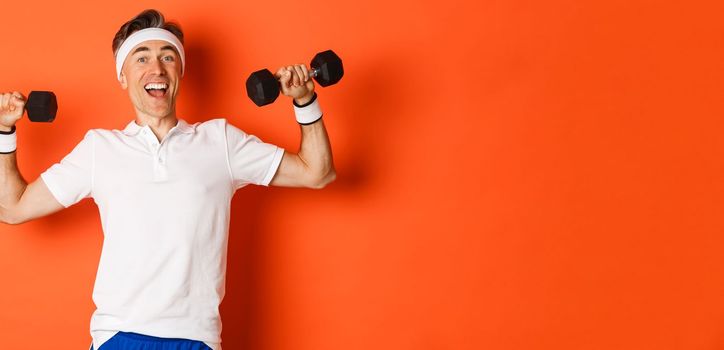 Concept of workout, gym and lifestyle. Image of healthy and strong middle-aged male athlete, doing sport exercises with dumbbells and smiling, standing over orange background.