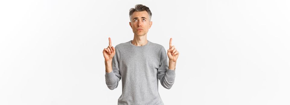 Portrait of gloomy and hesitant middle-aged man, looking troubled and pointing fingers up, standing upset over white background.