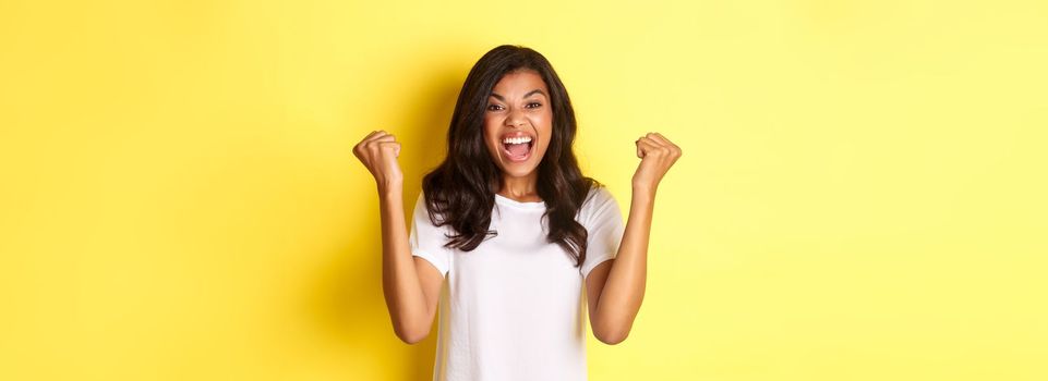 Image of successful african-american girl, feeling lucky, making fist pump sign and saying yes, triumphing and shouting for joy, standing over yellow background.