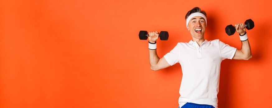 Concept of workout, gym and lifestyle. Image of healthy and strong middle-aged male athlete, doing sport exercises with dumbbells and smiling, standing over orange background.