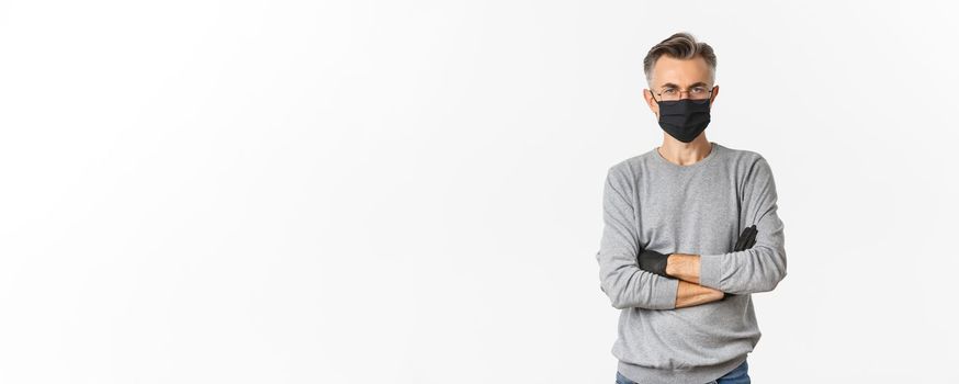 Concept of coronavirus, lifestyle and quarantine. Portrait of displeased middle-aged man in medical mask and gloves, cross arms on chest and looking disappointed, standing over white background.