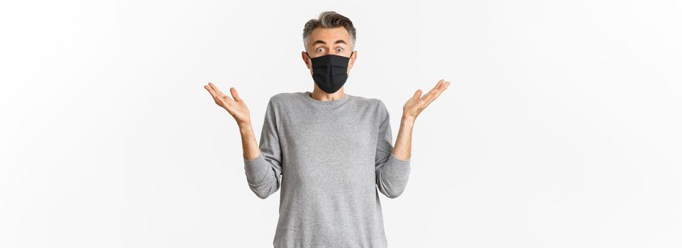 Covid-19, pandemic and social distancing concept. Portrait of handsome middle-aged man in black medical mask looking surprised, reacting amazed, standing over white background.