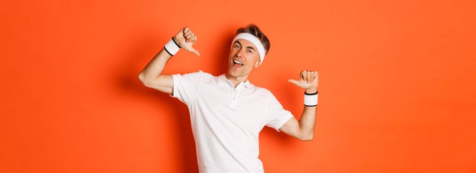 Concept of sport, fitness and lifestyle. Image of confident middle-aged man pointing at himself proudly, wearing clothes for workout, standing over orange background.