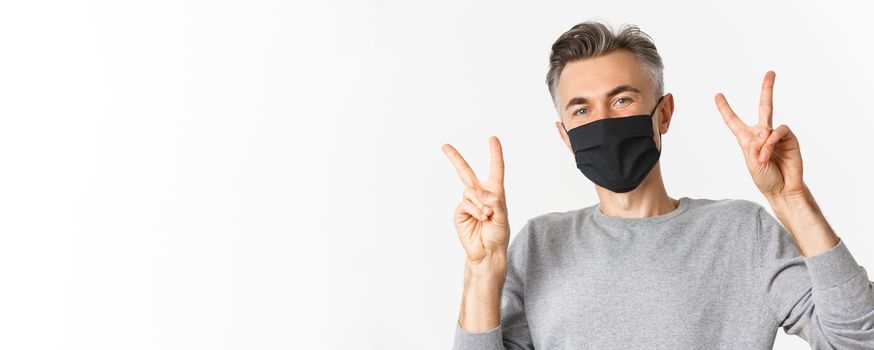 Concept of covid-19, social distancing and quarantine. Close-up of happy and cute middle-aged guy in black medical mask, showing peace signs and smiling, standing over white background.