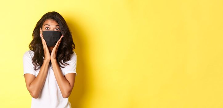 Concept of coronavirus, pandemic and lifestyle. Portrait of excited african-american woman in face mask, looking amazed at something cool, standing over yellow background.