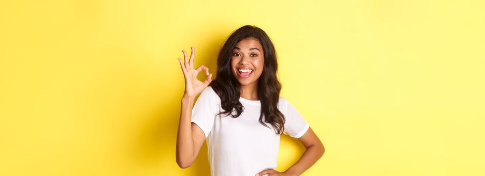 Portrait of happy good-looking african american girl, showing okay sign and smiling in approval, praise good work, standing over yellow background.