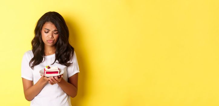 Image of sad african-american girl looking at piece of cake, cant eat it, being on diet, standing over yellow background.