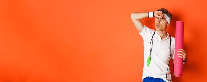 Concept of workout, gym and lifestyle. Image of handsome middle-aged man, tired after fitness exercises, holding skipping rope and yoga mat, wiping sweat off forehead, orange background.