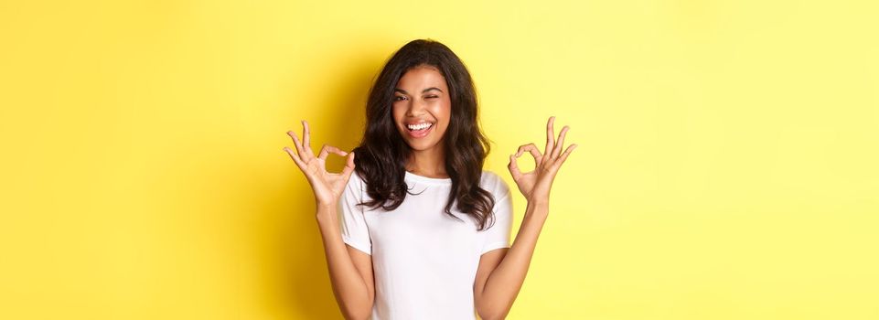 Portrait of confident, attractive african-american girl, showing okay signs and winking, guarantee quality, approve and agree something good, standing over yellow background.