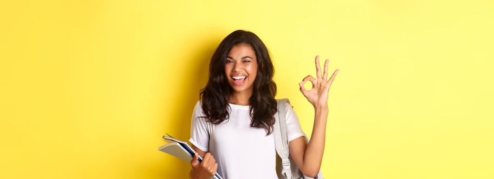 Portrait of confident african-american female student, showing okay sign and winking, approve something good, standing with notebooks and backpack for college, yellow background.