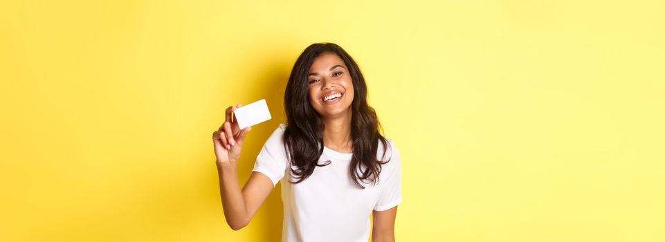 Image of lovely african-american woman smiling happy, showing credit card, standing over yellow background.