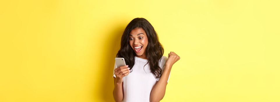 Portrait of beautiful african-american girl, rejoicing and looking at mobile phone, found something amazing online, standing over yellow background.