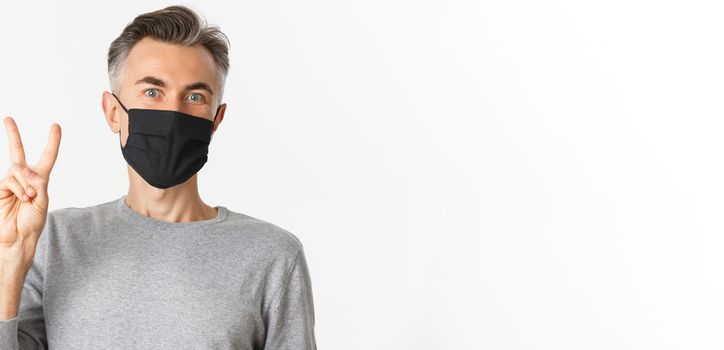 Concept of covid-19, social distancing and quarantine. Close-up of cheerful middle-aged guy in medical mask, showing peace sign and smiling, standing over white background.