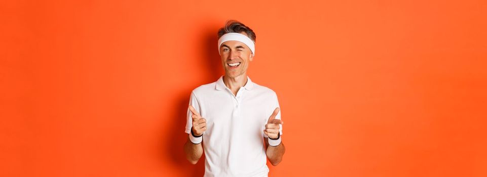 Concept of sport, fitness and lifestyle. Portrait of confident, handsome middle-aged man in workout uniform, pointing fingers at camera and smiling, inviting to gym, standing over orange background.