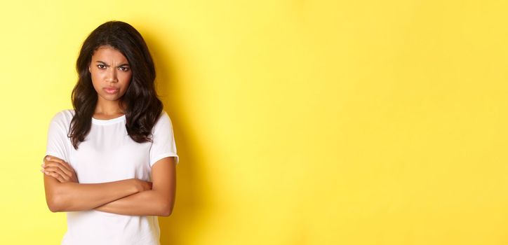 Image of cute african-american girl feeling angry, cross arms on chest and frowning, mad at you, standing over yellow background.