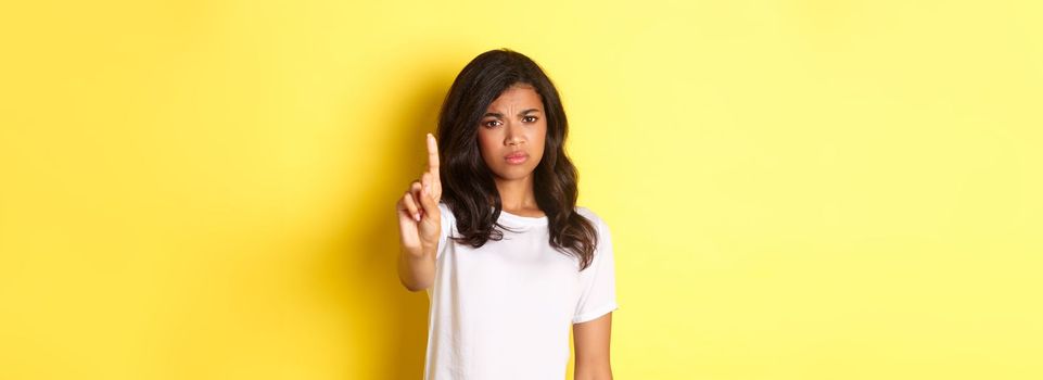 Image of disappointed african-american girl telling no, shaking finger to prohibit or stop someone, disagree with person, standing over yellow background.