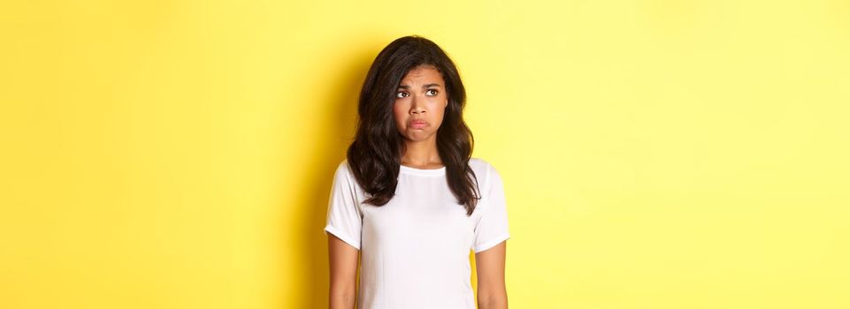 Portrait of sad and disappointed african-american woman, sulking and frowning as looking at upper left corner, see something upsetting, standing over yellow background.