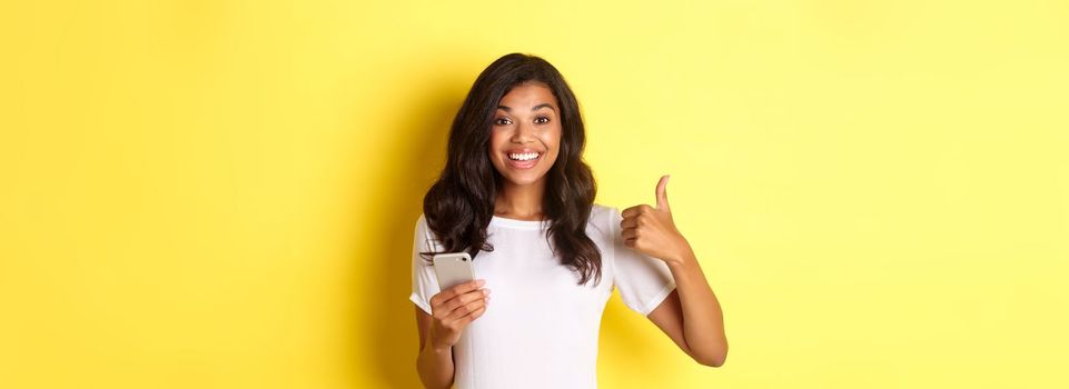 Portrait of young good-looking african american woman, showing thumbs-up while using mobile phone app, smiling pleased, standing over yellow background.