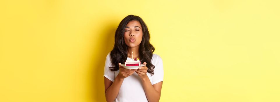 Portrait of beautiful african-american girl celebrating birthday, blowing lit candle on b-day cake and making a wish, standing over yellow background.