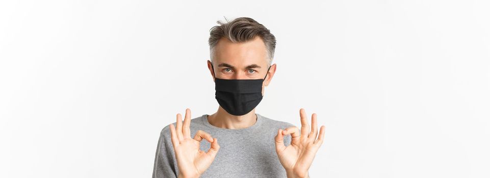 Concept of covid-19, social distancing and quarantine. Close-up of confident middle-aged man, wearing black medical mask, assure everything good, showing okay signs, white background.