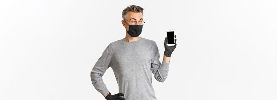 Concept of covid-19, social distancing and lifestyle. Portrait of handsome middle-aged man in medical mask, gloves and glasses, showing mobile phone screen, standing over white background.