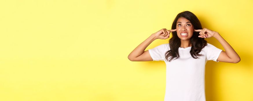 Image of annoyed and bothered african-american girl, shut ears and looking angry up, complaining at loud neighbours, standing over yellow background.