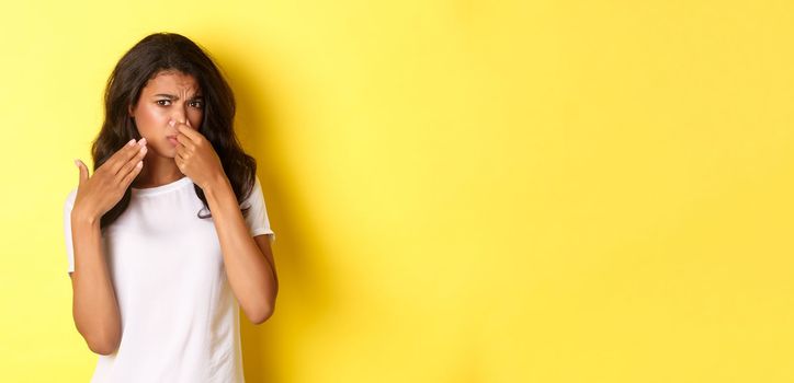 Portrait of young african-american girl, shut her nose and looking at something with disgusting smell, complaining on bad stink, standing over yellow background.