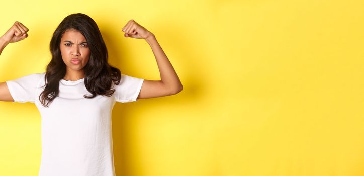 Image of strong and confident african-american girl, flexing biceps and looking self-assured, standing over yellow background.