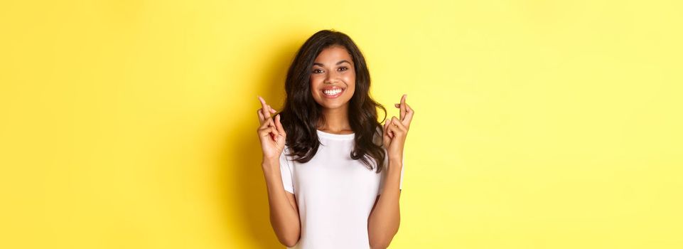 Portrait of optimistic, smiling african-american girl, crossing fingers for good luck and making wish, standing over yellow background.