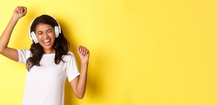 Cheerful teenage african-american girl listening to music in headphones, dancing upbeat and smiling, standing over yellow background.